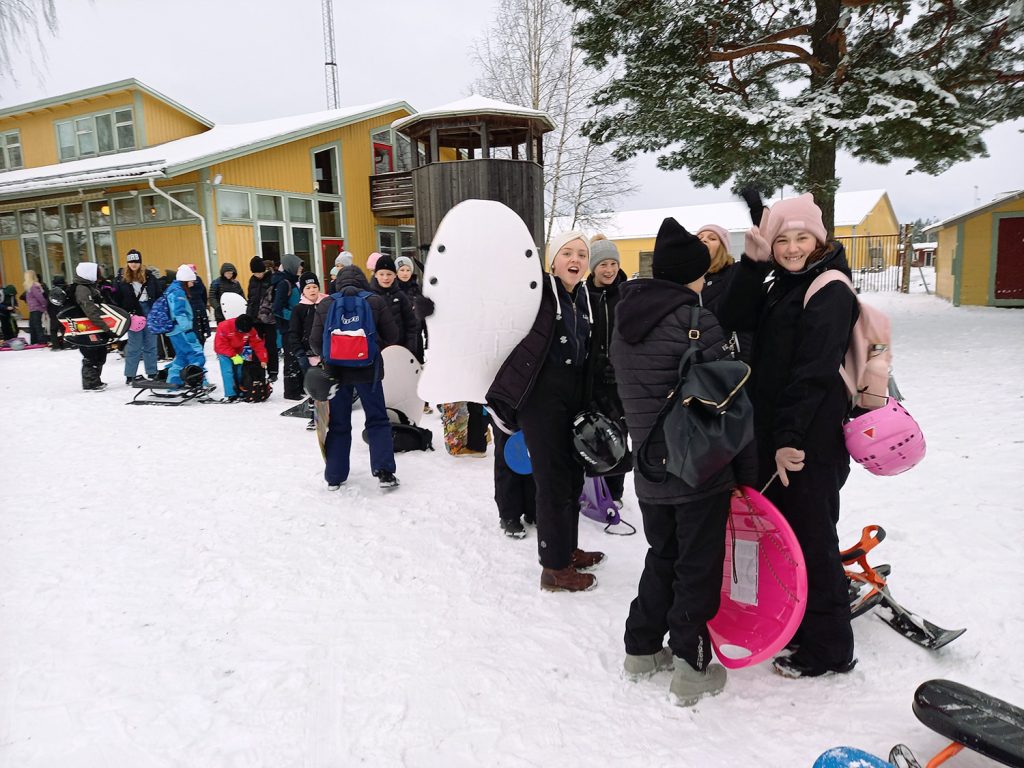 Barn som står på led med pulkor i snön framför en gul byggnad och några poserar för kameran.